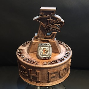 May include: A wooden trophy with a carved eagle emblem and the text "CHIEFS" on the base. A championship ring with the text "PATRIOT LEAGUE CHAMPIONS" and "BACK TO BACK" is displayed on the trophy. The years 2016, 2017, and 2018 are also visible.