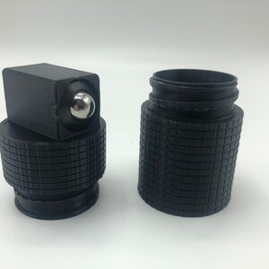 May include: A black, 3D-printed roller bottle with a textured grip. The bottle has a square top with a silver rollerball and a separate, textured base. The bottle is designed to hold essential oils or other liquids.