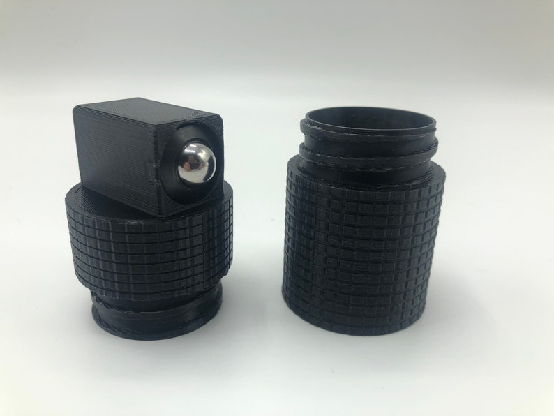 May include: A black, 3D-printed roller bottle with a textured grip. The bottle has a square top with a silver rollerball and a separate, textured base. The bottle is designed to hold essential oils or other liquids.