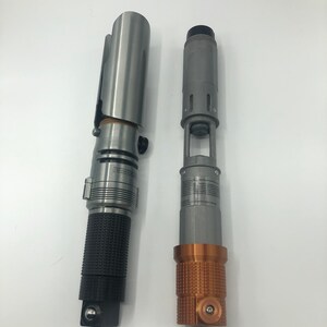 May include: Two metallic lightsaber props. One is silver with a black textured grip, the other is gray with a copper-colored textured base. Both have detailed designs and appear to be replicas or models.