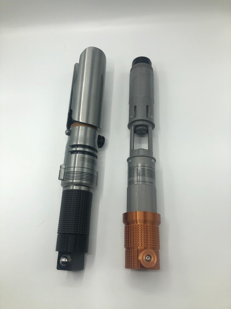 May include: Two metallic lightsaber props. One is silver with a black textured grip, the other is gray with a copper-colored textured base. Both have detailed designs and appear to be replicas or models.