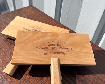 May include: Two wooden Howard Brush Company paint paddles with handles. The paddles are rectangular with rounded corners and feature the company's logo and text. The paddles are made in the USA.