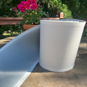 May include: A roll of light gray, textured material with a repeating diagonal pattern. The material is unrolled, revealing a flat surface with the same texture. A potted plant with bright pink flowers is in the background.