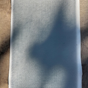 May include: A rectangular piece of light gray fabric with a textured surface. The edges are finished with a thin black border. The fabric appears to be a woven material, possibly for upholstery or crafting.
