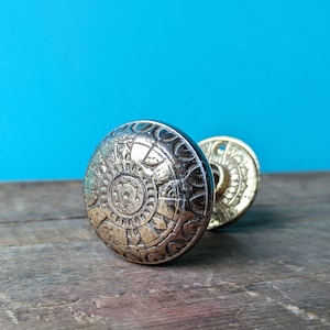May include: A vintage brass door knob with a decorative, circular design. The knob is on a wooden surface.