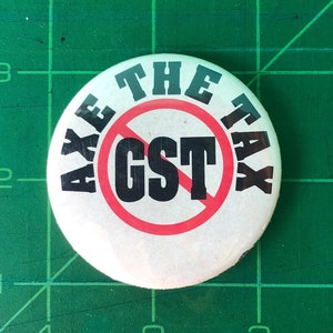 Peut inclure: Un bouton blanc avec un cercle rouge et une ligne diagonale qui le traverse. Le texte "AXE THE TAX" est écrit autour du cercle. À l'intérieur du cercle, le texte "GST" est écrit en noir.