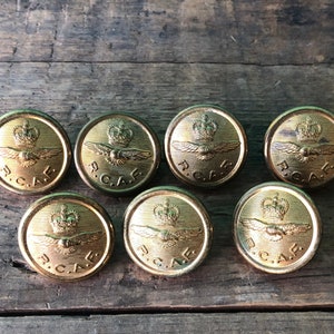 May include: Eight gold-coloured metal buttons featuring the Royal Canadian Air Force (R.C.A.F.) logo and a crown on top. The buttons are arranged in two rows of four on a wooden surface.