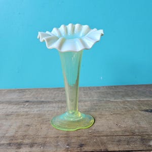 Op de afbeelding: Een lichtgele glazen vaas met een gerimpelde, witte rand. De vaas heeft een slanke, langwerpige body en een cirkelvormige basis. De achtergrond is effen turquoise en de vaas staat op een verweerd houten oppervlak.