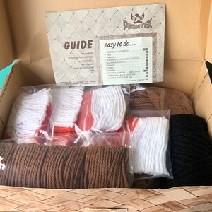 May include: A box filled with craft supplies for a rug hooking project. The box contains bags of yarn in brown, orange, white, and black. The box also includes a guide with instructions on how to do rug hooking. The guide has the text "GUIDE" and "easy to do..." on it.