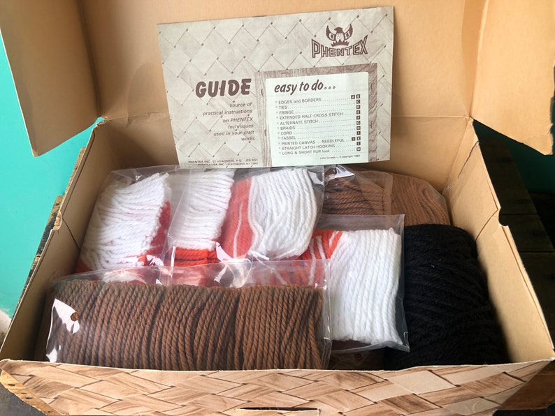 May include: A box filled with craft supplies for a rug hooking project. The box contains bags of yarn in brown, orange, white, and black. The box also includes a guide with instructions on how to do rug hooking. The guide has the text "GUIDE" and "easy to do..." on it.