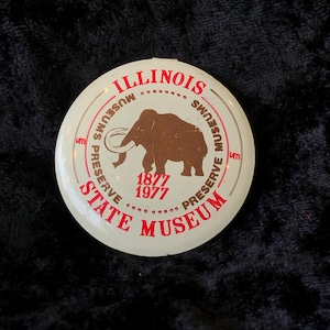 Peut inclure: Un bouton blanc avec du texte rouge qui dit "Illinois State Museum Preserve Museums 1877 1977 Preserve". Le bouton présente une silhouette brune d'un mammouth.