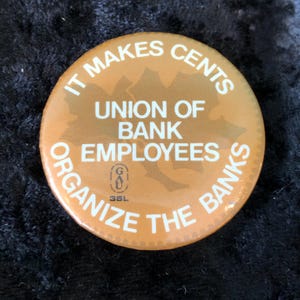 Könnte beinhalten: Runder, orangefarbener Button mit weißem Text. Der Text lautet "IT MAKES CENTS", "UNION OF BANK EMPLOYEES" und "ORGANIZE THE BANKS". In der Mitte befindet sich eine Berg-Grafik. Vintage-Artikel.