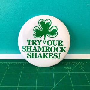 Könnte beinhalten: Ein weißer Knopf mit einem grünen Kleeblatt und dem Text "TRY OUR SHAMROCK SHAKES!". Der Knopf ist rund und hat eine glänzende Oberfläche. Der Hintergrund ist eine türkisfarbene Wand und eine grüne Schneidematte.