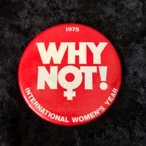 Könnte beinhalten: Ein runder, roter Button mit weißem Text "WHY NOT!" und einem weiblichen Symbol. Der Text "1975" steht oben, und "INTERNATIONAL WOMEN'S YEAR" steht am Rand.