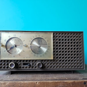 May include: A vintage brown and gold clock radio with a black speaker grille. The radio has a clock face with black hands and a dial for tuning AM and FM stations. The radio is sitting on a wooden surface.