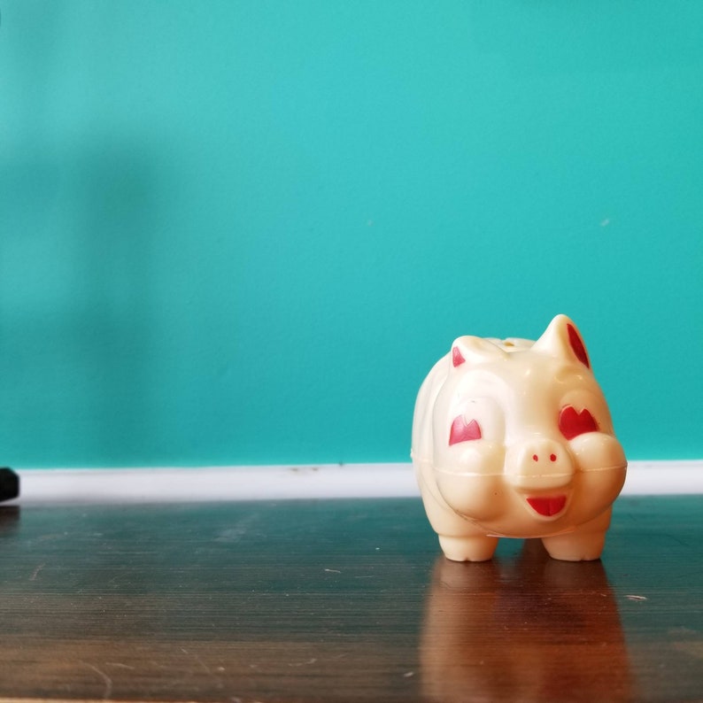 May include: A white plastic piggy bank with red painted eyes and a smiling mouth. The piggy bank is sitting on a dark wood surface.