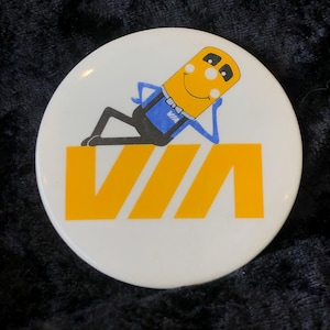 May include: A white button featuring a yellow and black logo for VIA. The logo depicts a cartoon stick figure with a yellow body, blue overalls, and a yellow bow tie. The stick figure is leaning back with a smile on its face.