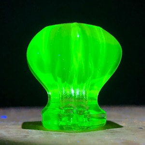 May include: A bright, luminous green glass knob with a rounded top and a flared base. The knob has a ribbed texture on the top section and sits on a surface. The background is dark.