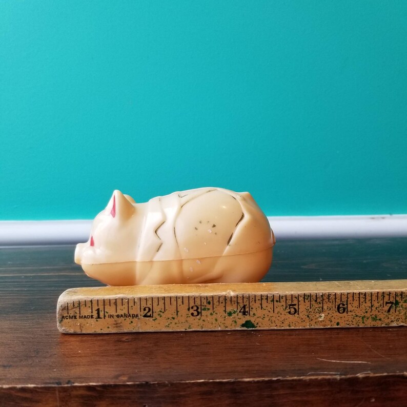 May include: A white plastic piggy bank with red accents. The piggy bank is lying on its side and measures approximately 10 cm long. The words "ACME MADE IN CANADA" are printed on the wooden ruler behind the piggy bank.