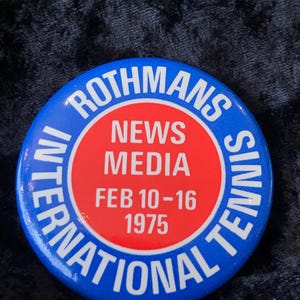 Könnte beinhalten: Ein runder, blauer und roter Button mit der Aufschrift "ROTHMANS INTERNATIONAL TENNIS NEWS MEDIA" und "FEB 10-16 1975". Der Button hat eine glänzende Oberfläche und einen Vintage-Look.