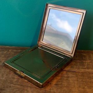 May include: A gold-toned compact mirror with a hinged lid and a circular patterned design on the inside. The mirror is open and the reflection of a cloudy sky is visible.