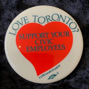 Könnte beinhalten: Ein weißer Knopf mit einem roten Herzen in der Mitte. Das Herz enthält den Text "Support Your Civic Employees". Der Knopf hat auch den Text "Love Toronto?" oben und "CUPE Locals 43 and 79" unten.
