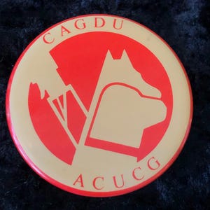 Könnte beinhalten: Ein runder Knopf in Rot und Beige mit dem Text "CAGDU ACUCC" in weißen Buchstaben. Der Knopf zeigt eine stilisierte Silhouette eines Hundekopfes.