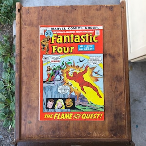 Può includere: Una copertina di fumetto vintage con i Fantastici Quattro. La copertina è rossa, gialla e blu con una grande immagine della Torcia Umana in volo. Il testo sulla copertina recita "Fantastic Four" e "The Flame and the Quest!"