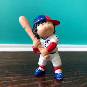 Può includere: Piccola statuetta di un giocatore di baseball in uniforme bianca con dettagli rossi e blu. Il giocatore tiene in mano una mazza di legno e indossa un berretto blu e rosso. La statuetta è su una superficie di legno scuro.