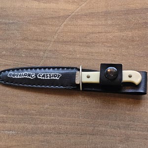 May include: A small knife with a cream-colored handle and a silver blade is sheathed in a black leather case. The case is secured with a black leather strap and a metal stud. The text "HOPALONG CASSIDY" is printed on the case.
