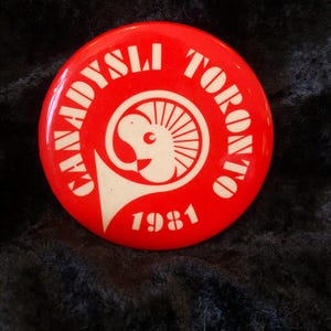 Könnte beinhalten: Ein roter Knopf mit weißem Text und einer Grafik. Der Text lautet "Canadysli Toronto 1981". Die Grafik ist ein stilisierter Widderkopf mit einer Trompete. Der Knopf ist kreisförmig und hat ein glänzendes Finish.