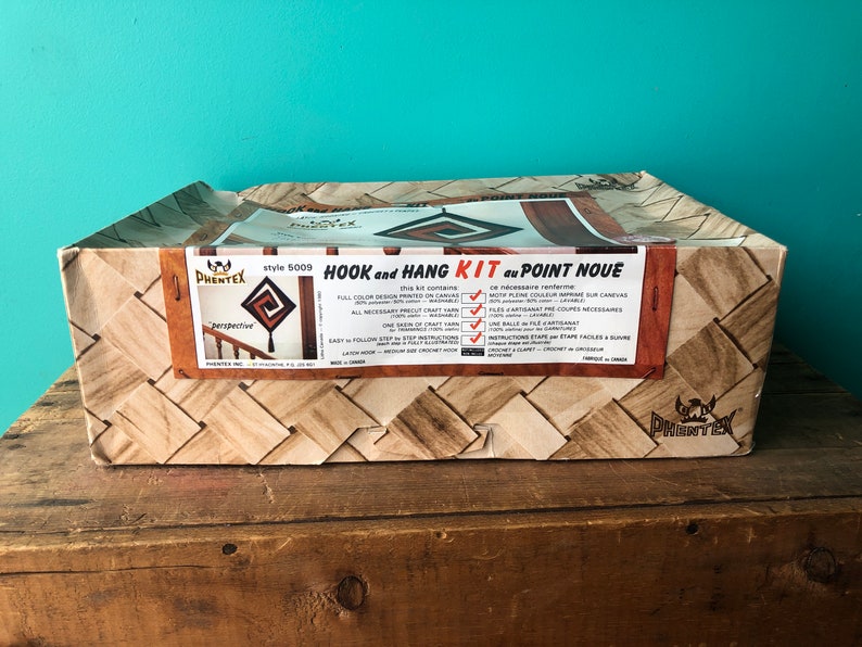 May include: A brown cardboard box with a woven pattern. The box contains a "Hook and Hang Kit du Point Noue" craft kit. The kit includes all the materials needed to create a wall hanging. The box features a picture of the finished product, a geometric design with a spiral pattern. The text on the box reads "Style 5009" and "Perspective".