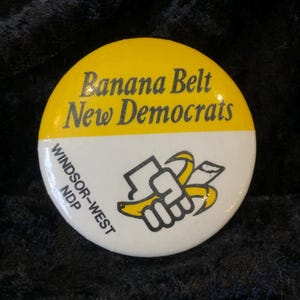 May include: A circular badge with a yellow upper section and a white lower section. The yellow part has the text "Banana Belt New Democrats". The white section features a fist holding a peeled banana and the text "Windsor-West NDP".
