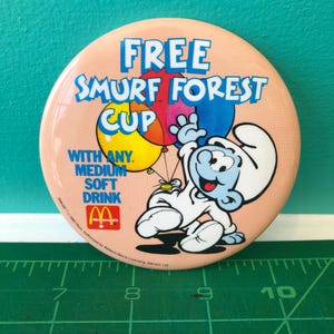 Könnte beinhalten: Ein runder, pfirsichfarbener Button mit einem Schlumpf, der Luftballons hält. Der Button trägt den Text "FREE SMURF FOREST CUP" und "WITH ANY MEDIUM SOFT DRINK" sowie das McDonald's Logo.