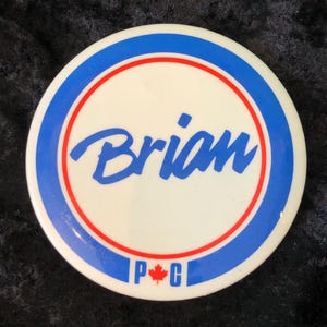 Könnte beinhalten: Ein weißer runder Knopf mit einem blauen und roten Rand. Der Knopf trägt den Namen "Brian" in blauer Kursivschrift und die Buchstaben "P.C." mit einem roten Ahornblatt dazwischen.