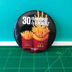 Könnte beinhalten: Ein runder, schwarzer Button mit dem Text "30% MORE FRIES!" und zwei McDonald's Pommes-Frites-Behältern, gefüllt mit goldenen Pommes. Das McDonald's-Logo ist auf den Behältern zu sehen. Der Text "APPROX. BY VOLUME." ist ebenfalls vorhanden.