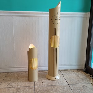 May include: Two tall, cream-coloured lamps with a cylindrical design. The larger lamp has a clock face and a circular cutout. The smaller lamp has a rectangular cutout. Both lamps have a light source inside.