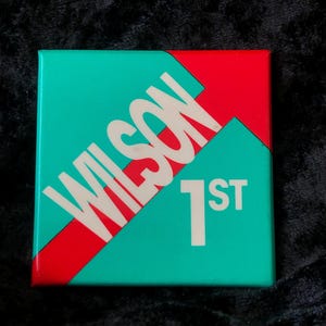 Könnte beinhalten: Ein quadratischer Knopf in Türkisblau und Rot mit dem Wort "WILSON" in weißen Buchstaben und der Zahl "1ST" in weißen Buchstaben.