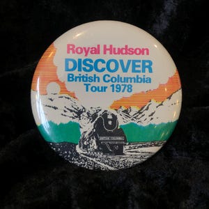 Könnte beinhalten: Ein weißer Knopf mit einem bunten Bild eines Zuges, der durch eine Berglandschaft fährt. Der Text "Royal Hudson Discover British Columbia Tour 1978" ist auf dem Knopf gedruckt.