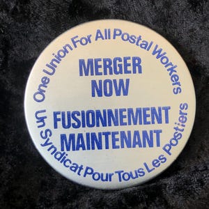 Könnte beinhalten: Ein weißer Knopf mit blauem Text, der "One Union For All Postal Workers" und "Un Syndicat Pour Tous Les Postiers" lautet. Der Knopf hat auch den Text "Merger Now" und "Fusionnement Maintenant".