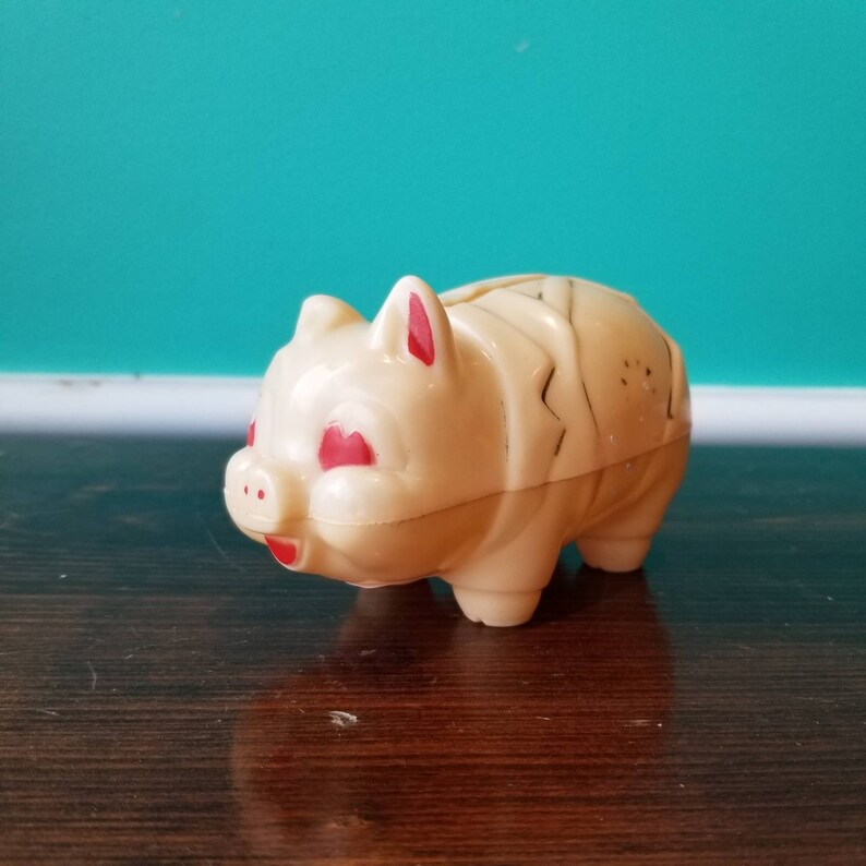 May include: A vintage plastic piggy bank with a cream-coloured body and red painted eyes and ears. The piggy bank has a slot on the top for coins.