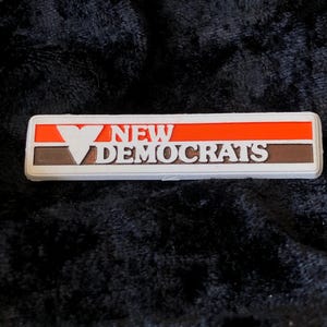 Könnte beinhalten: Ein weißer rechteckiger Knopf mit einem roten und braunen Streifen. Der Knopf hat ein weißes Herz mit einem weißen Umriss und den Text "NEW DEMOCRATS" in Weiß.