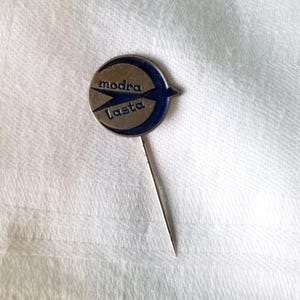 May include: A vintage pin with a circular design featuring the words "modra lasta" in a stylized font. The pin has a blue border and a silver pin backing. The pin is set against a white fabric background.
