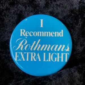 Könnte beinhalten: Ein runder, hellblauer Button mit weißem Text. Der Text lautet "I Recommend Rothmans EXTRA LIGHT". Es ist ein Vintage-Werbeartikel.