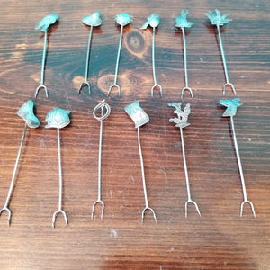 Op de afbeelding: Een verzameling van elf zilverkleurige cocktailprikkers met decoratieve toppen. De toppen hebben verschillende ontwerpen, waaronder vogels, hoeden en abstracte vormen. Elke prikker heeft een lange, slanke steel en een vork met twee tanden aan de onderkant.