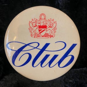 Könnte beinhalten: Ein weißer runder Knopf mit einem roten und blauen Wappen und dem Wort "Club" in blauer Kursivschrift.