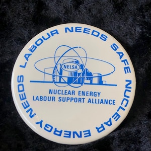 May include: White button with blue text and a graphic of a nuclear power plant. The text reads "Labour Needs Safe Nuclear Energy", "Nuclear Energy Labour Support Alliance", and "NELSA".