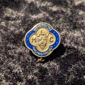 May include: A gold and blue enamel pin with the text "Universitas McMaster" and the letters "LMC" in the center. The pin features a crest with a building and a maple leaf.