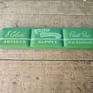 Peut inclure: Une boîte de peinture vintage en métal vert avec le texte "8 Colour Artists" à gauche, "Asco Water Colours Supply" au milieu et "Paint Box Co. Limited" à droite.