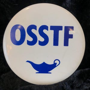 Könnte beinhalten: Ein weißer Knopf mit dem Text "OSSTF" in blauen Buchstaben und einer blauen Öllampengrafik.
